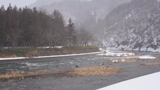 福島県南会津町の伊南川、雪が降る残雪の川岸と冬の山林を流れる清流4K映像