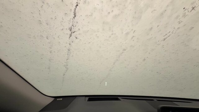 Foam and water drops flowing over car windshield during automatic car wash. View from inside vehicle through wet glass with bubbles and blurred lights outside.