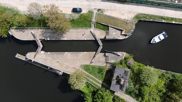 Aerial view of a canal lock system with a small white boat navigating the dark water surrounded by green trees and paths in Knottingley, England, United Kingdom.