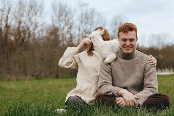 Naklejka premium A couple enjoys an outdoor moment with a playful goat kid, sharing affection and laughter, sitting on grass in a park, showcasing a casual lifestyle and warm bond