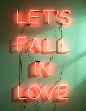 Neon sign spelling "LET'S FALL IN LOVE" on a teal wall, casting shadows and illuminated by green light filtering from the side.