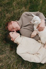Naklejka premium Relaxed couple lying on the grass, sharing a gentle moment with a white goat kid. Soft natural light, cozy sweaters, outdoor scenery evokes wholesome lifestyle and bonding and shared joy.