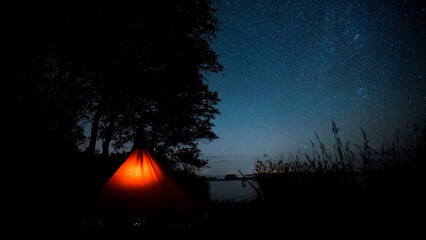 Starry night sky over a tranquil lake, an illuminated orange tent glowing under the vast dark cosmos, serene outdoor camping adventure, peaceful wilderness escape. © Shanza