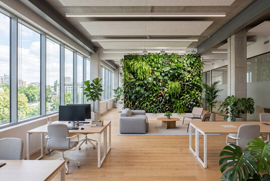 Open space luminoso con parete verde, grandi finestre, postazioni di lavoro e area lounge. Scena che rappresenta design biophilico, benessere in ufficio e ambiente di lavoro moderno.