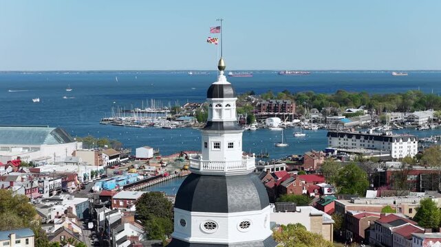 Aerial close orbit of Maryland State House dome in Annapolis, Maryland with USA and state flags waving above harbor, marina and Chesapeake Bay on sunny winds spring day with historic downtown skyline.
