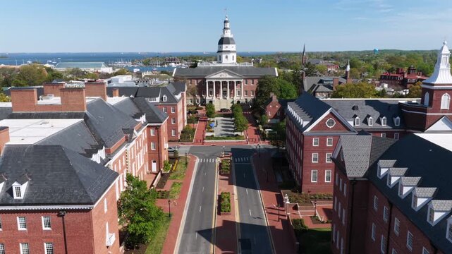 Aerial approach to Maryland State House in Annapolis, Maryland framed by red brick buildings and downtown streets, with Chesapeake Bay beyond under bright spring sunshine in historic USA capital city.