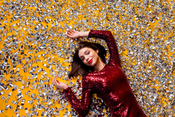 Naklejka premium Woman in red sequin dress lying in silver confetti on yellow background celebrating glamour and joy