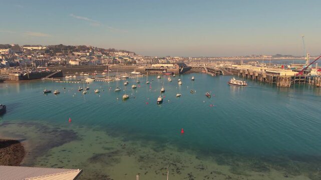 St Peter Port Harbour Guernsey.Low drone footage across the outer harbour to the ferry berth with boats on swi8nging moorings and small ferry arriving on bright sunny afternoon with clear blue water