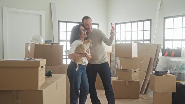 Man and woman holding small dog while man taps red smartphone screen among stacked moving boxes in building; new beginning joy.