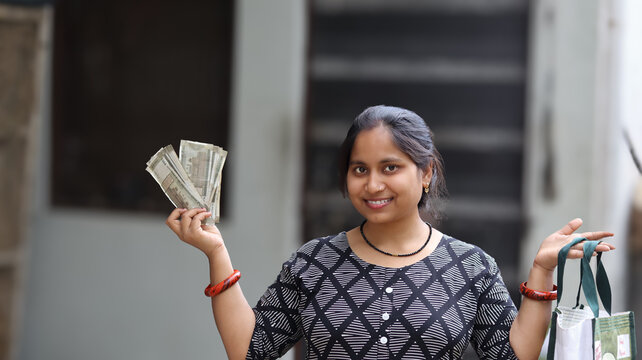 Happy Indian woman showing fan of five hundred rupee notes and holding shopping bag, cheerful female consumer with cash and groceries, financial freedom concept
