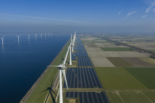 Aerial view of a massive renewable energy park featuring a long row of wind turbines and extensive solar panel arrays along a coastal dike under a clear blue sky.