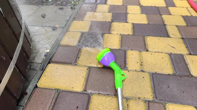Colorful garden sprayer lies on stone pavement outdoor. Garden sprayer waters backyard tiles with fresh liquid. Plastic nozzle irrigation tool helps cleaning path or watering garden area.