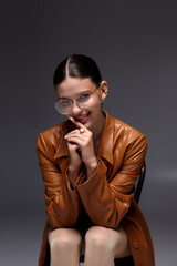 Stylish young woman in leather coat and glasses poses elegantly in studio setting
