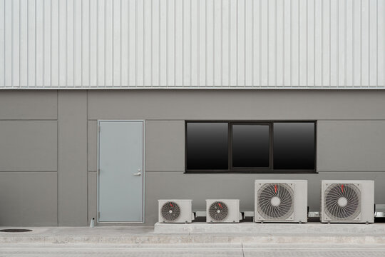 Modern industrial building exterior featuring a grey metal door, dark tinted windows, and four air conditioning compressor units on ground.