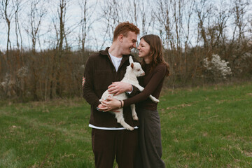 Naklejka premium Cheerful couple outdoors, embracing a goat kid, smiling together in a sunlit field, calm countryside mood, natural colors, relaxed posture, tender connection, casual fashion