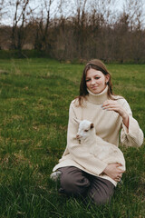 Naklejka premium A woman in a cozy sweater sits in a grassy field, gently holding a young goat kid on her lap. A calm lifestyle moment capturing bond with animals and open outdoors.