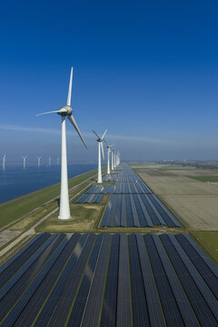 Aerial view of a large solar farm and a row of wind turbines along a coastal dike under a clear blue sky in Creil, Flevoland, Netherlands.