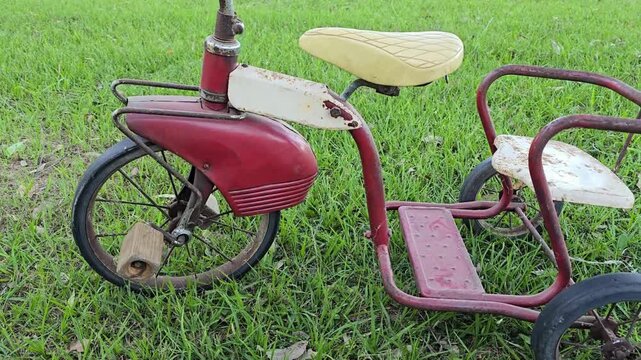 Vintage red tricycle with worn seat and rusted frame beside grass, captured in motion as camera pans across its aged metal body.