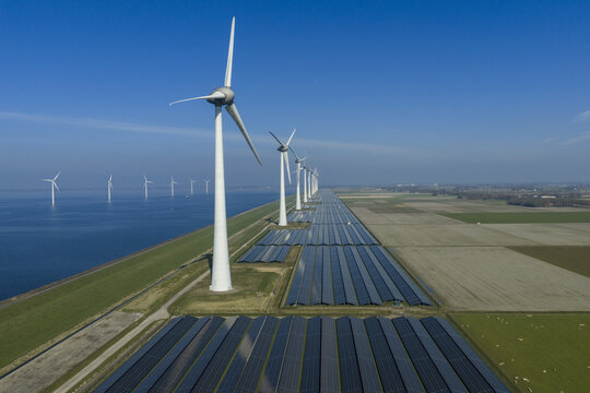 Aerial view of a row of modern wind turbines in the water and vast solar panel arrays on land stretching along a coastal dike under a clear blue sky in Netherlands.