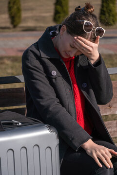 Woman sitting on an outdoor bench with a suitcase, covering her face with her hand due to a headache and feeling overwhelmed by travel stress and exhaustion. Vertical photo