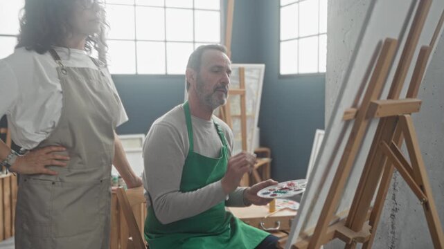 Man painter seated painting on easel holding palette while woman painter stands behind pointing hand in studio; creative collaboration concentration.
