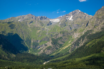 Obraz premium Expansive green valley opening to towering snow-patched mountain ranges under a clear blue sky, vibrant summer landscape ideal for hiking, trekking and outdoor adventure
