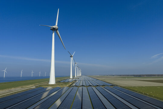 Aerial view of a large solar farm and a row of wind turbines along a coastal dike under a clear blue sky Netherlands.