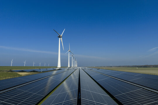 Aerial view of a large solar farm with rows of photovoltaic panels and tall wind turbines under a clear blue sky in Netherlands.
