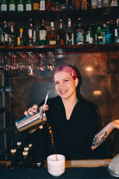 Smiling bartender with pink hair pouring cocktail at the bar