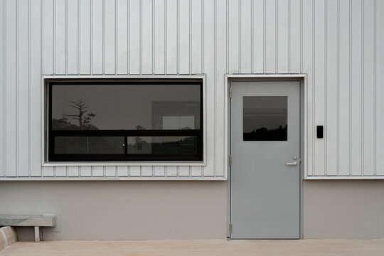 Exterior of modern industrial building with white corrugated metal wall, closed grey door, and dark tinted window reflecting trees.