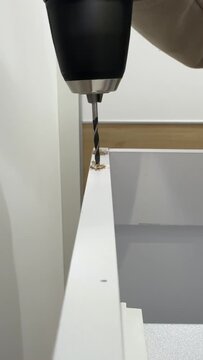 Hand using cordless drill on white furniture board edge with wood shavings during assembly. Vertical video footage for social media content and home improvement tutorials.