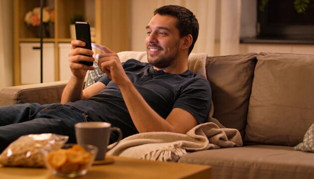 Reclining man in dark t-shirt and jeans, scrolling smartphone on beige sofa at home with snacks