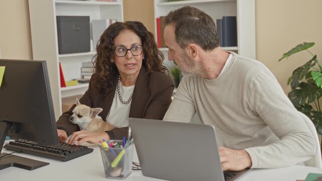 Middle aged woman holds small dog while man types on laptop at office desk, both reviewing screens together; focused teamwork companionship.