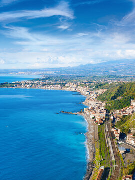 Aerial view of Taormina-Giardini Railway Station and Giardini Naxos coastline, Sicily, Italy. Mediterranean coastline stretches toward horizon under blue sky with wispy clouds