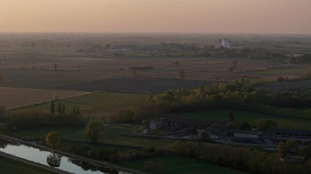Po Valley near Spinadesco Cremona Lombardy Italy with Canale Milano Cremona Po alongside cultivated fields rural buildings and distant grain silos under sunset haze across alluvial plain, aerial shot