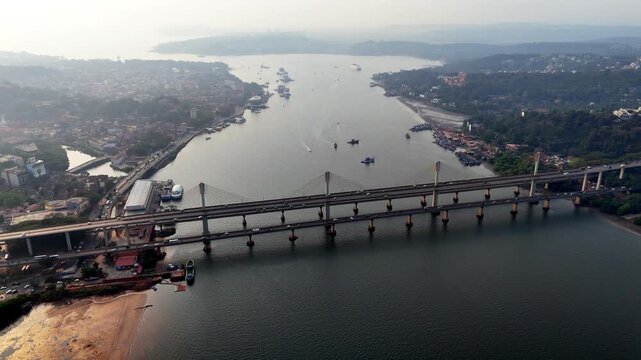 Cinematic aerial view of the Atal Setu spanning the Mandovi River, with boats scattered across calm waters and cityscapes stretching along both banks under a soft hazy sky.