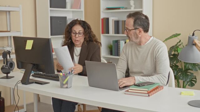Woman holding papers points and gestures with hand toward laptop on a desk in a studio office near monitor and pen cup while speaking to a seated man; frustration.