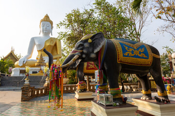 A magnificent large sitting Buddha statue with golden robes stands behind a colorful elephant statue in a beautiful Thai temple courtyard. © wannasak