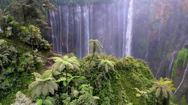 Aerial drone view of Tumpak Sewu waterfall in East Java, Indonesia. Spectacular curtain waterfall surrounded by dense tropical jungle and cliff edge with visible trail.