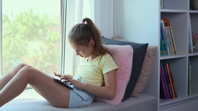 A little girl of European appearance is using a tablet. The kid is playing virtual games or watching cartoons on her tablet. The child with a tablet. The problem of addiction to gadgets.
