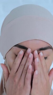 Close up of a young muslim woman wearing a traditional hijab, performing the wudu purification ritual by washing her face with water before praying, isolated on a white background