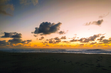 Fototapeta premium Experience tranquility with this stunning sunset over a dark sandy beach. Footprints symbolize a lonely journey across the vast ocean shore, perfect for wellness and travel marketing themes.