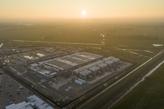 Aerial view of the Google data center facility with rows of cooling units and a parking lot surrounded by vast green fields and narrow canals under a hazy golden sunset.