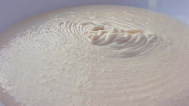 Creamy mixture flows smoothly into a white bowl, creating ripples and patterns on the surface as it settles, showcasing a continuous pouring action in a bright kitchen setting