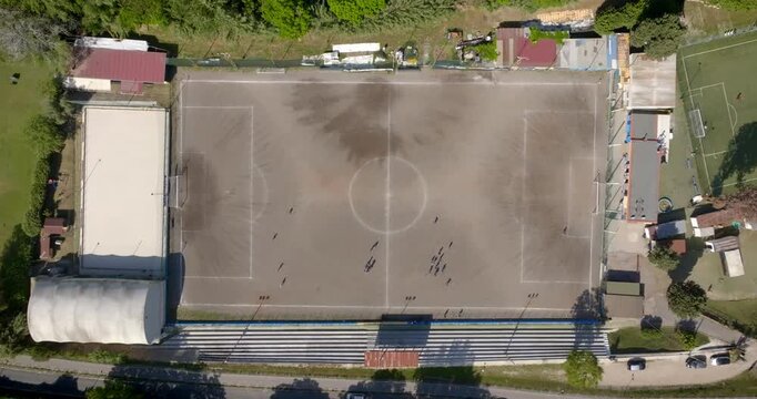 Top-down aerial view of a dirt soccer field featuring players, white boundary lines, and a central circle. The zenithal shot includes adjacent bleachers, facilities, and a sandy secondary court area.