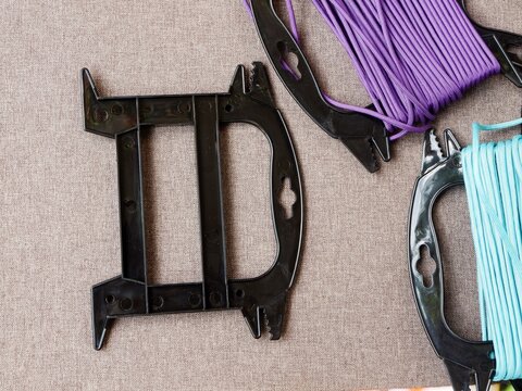 An empty black plastic cord winder alongside spools of purple and teal nylon paracord on a textured brown fabric surface.