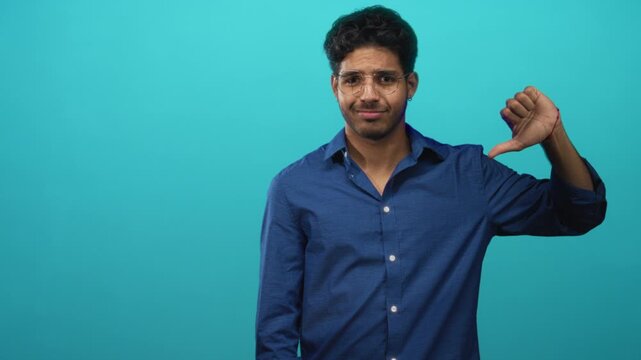 Man giving a thumbs down with bare thumb visible, eyes closed and glasses, wearing a blue button shirt against a teal studio backdrop; disapproval critique.