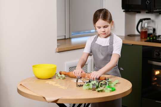 Little girl in apron rolling dough with wooden rolling pin, independent child cooking in the kitchen, early development of culinary skills and domestic lifestyle concept, making homemade cookies