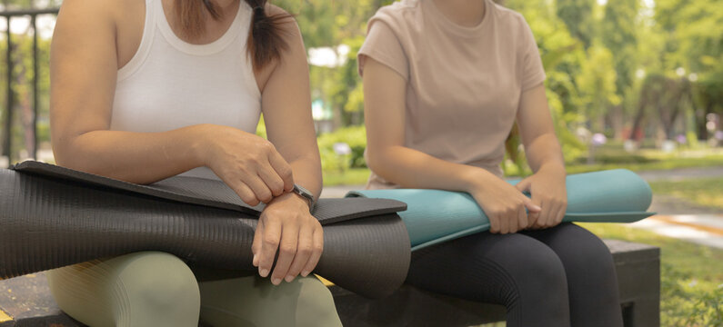 Two women sit on benches in a park, holding yoga mats. They seem to rest and prepare for their workout session in the morning sunlight. Nature surrounds them, creating a lively scene