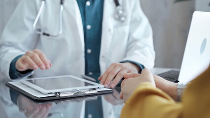 Fototapeta premium Doctor and patient consultation in clinic. Patient woman is gesturing over a glass desk while a physician is using tablet computer making some notes, close up of hands. Medicine and health care